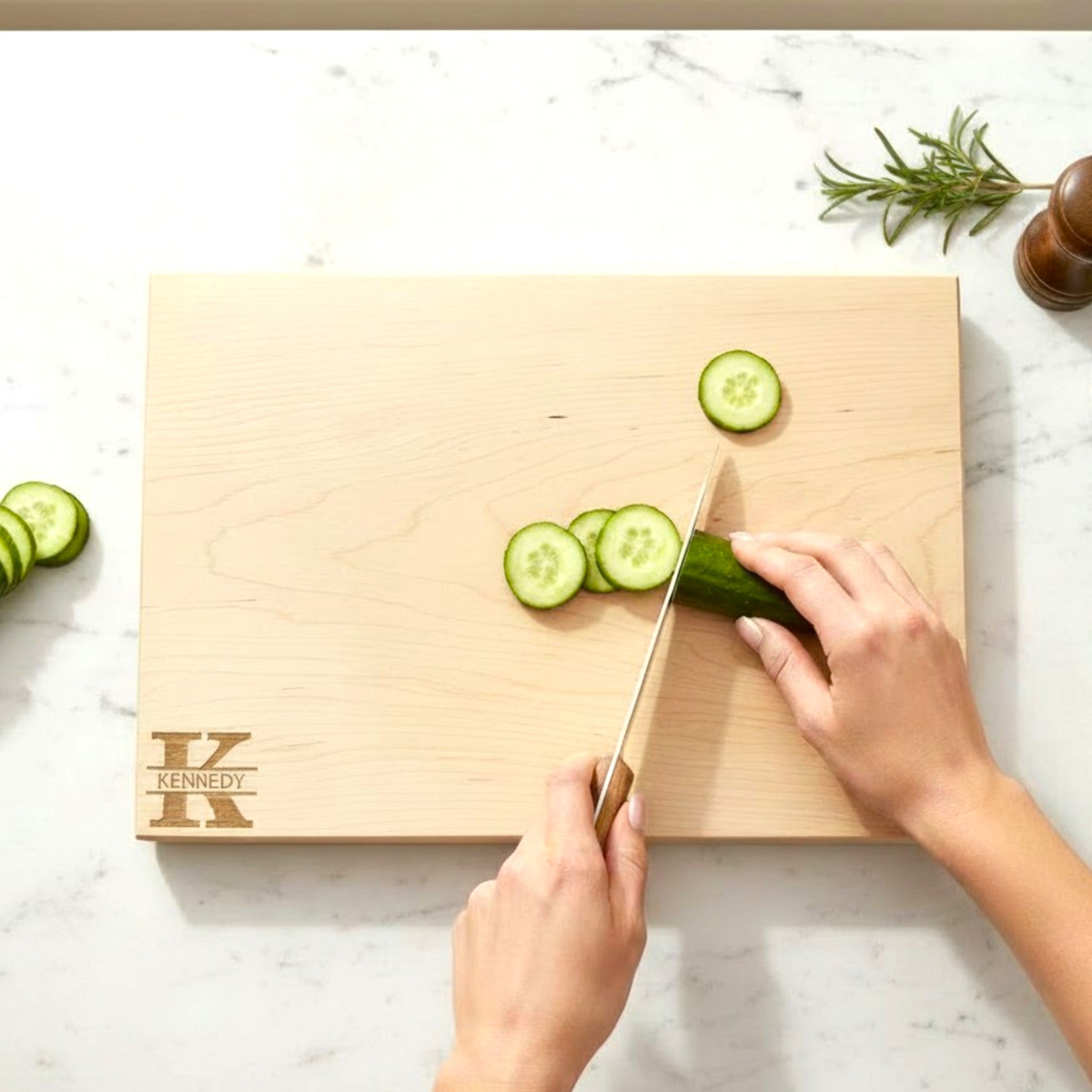 Person slicing cucumbers on a maple wooden cutting board with personalized laser engraved design and text on a white kitchen countertop.
